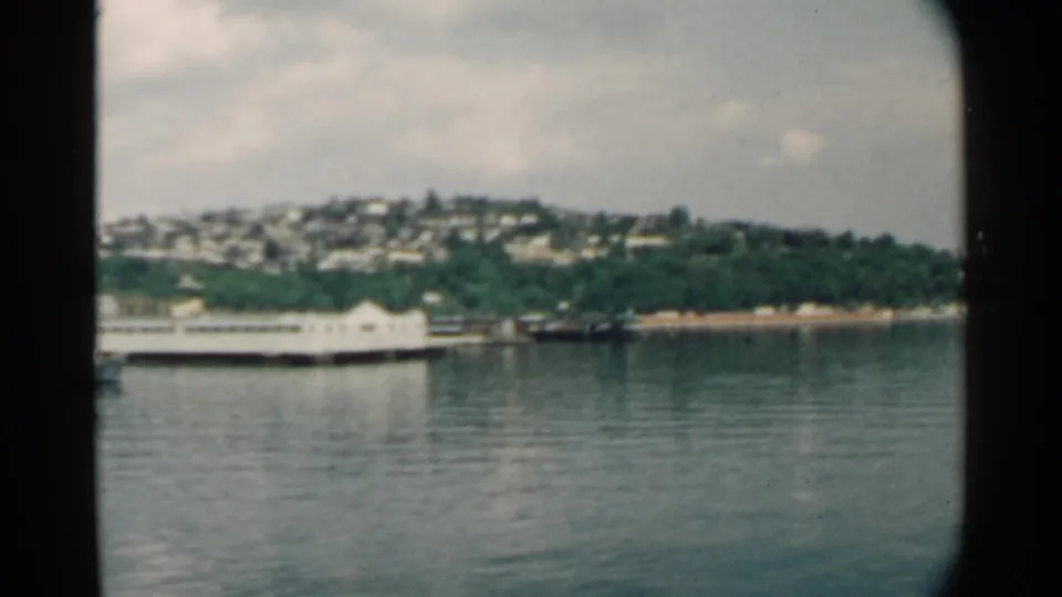 1958 Pearl Harbor view across calm water toward a long waterfront pier, green shoreline, and hillside housing under cloudy sky.