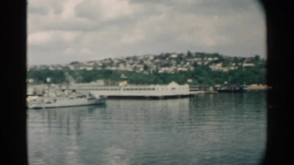 1958 Pearl Harbor view with boats near the waterfront, a long pier, reflective water, and hillside housing beyond the shoreline.