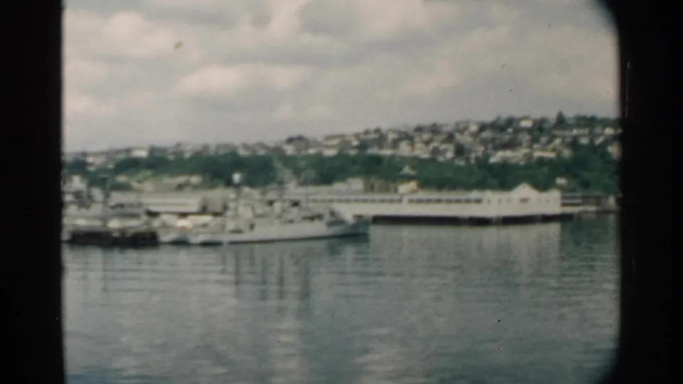 1958 Pearl Harbor frame showing a large white ship, smaller boats, a long pier, calm water, and hillside housing in the background.