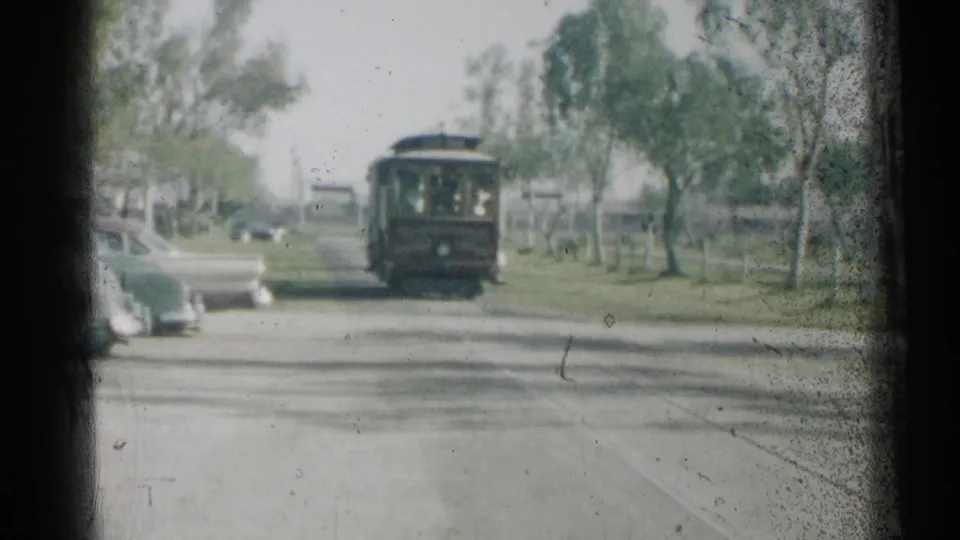 A red trolley-like vehicle approaches along a roadway, with parked mid-century cars, trees, grass, and dark film borders visible.