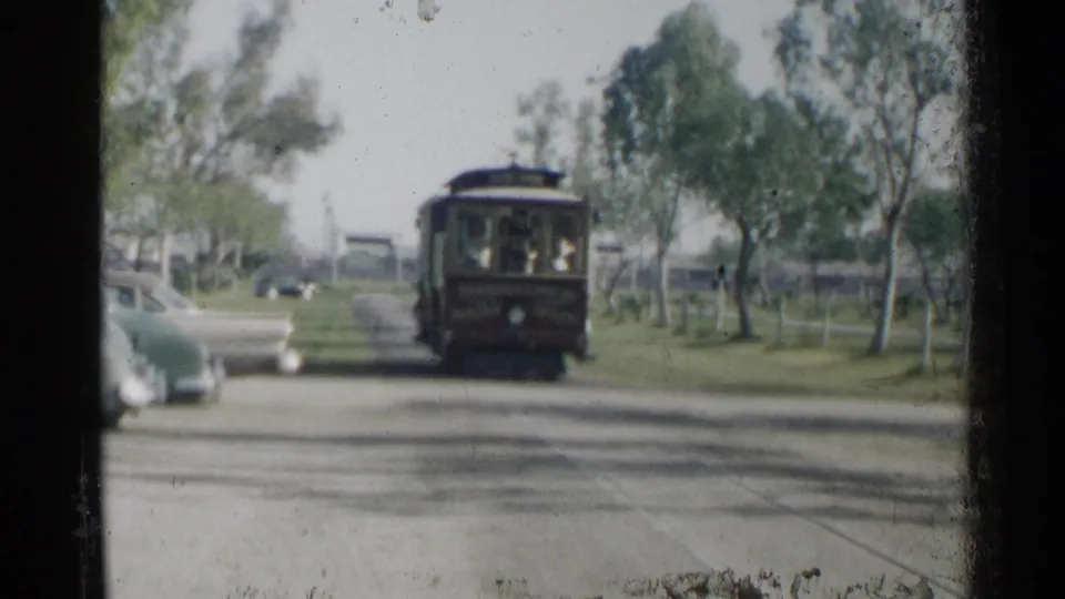 The red trolley-like vehicle appears closer in the frame, with passengers visible through front windows, parked cars nearby, and trees lining the road.