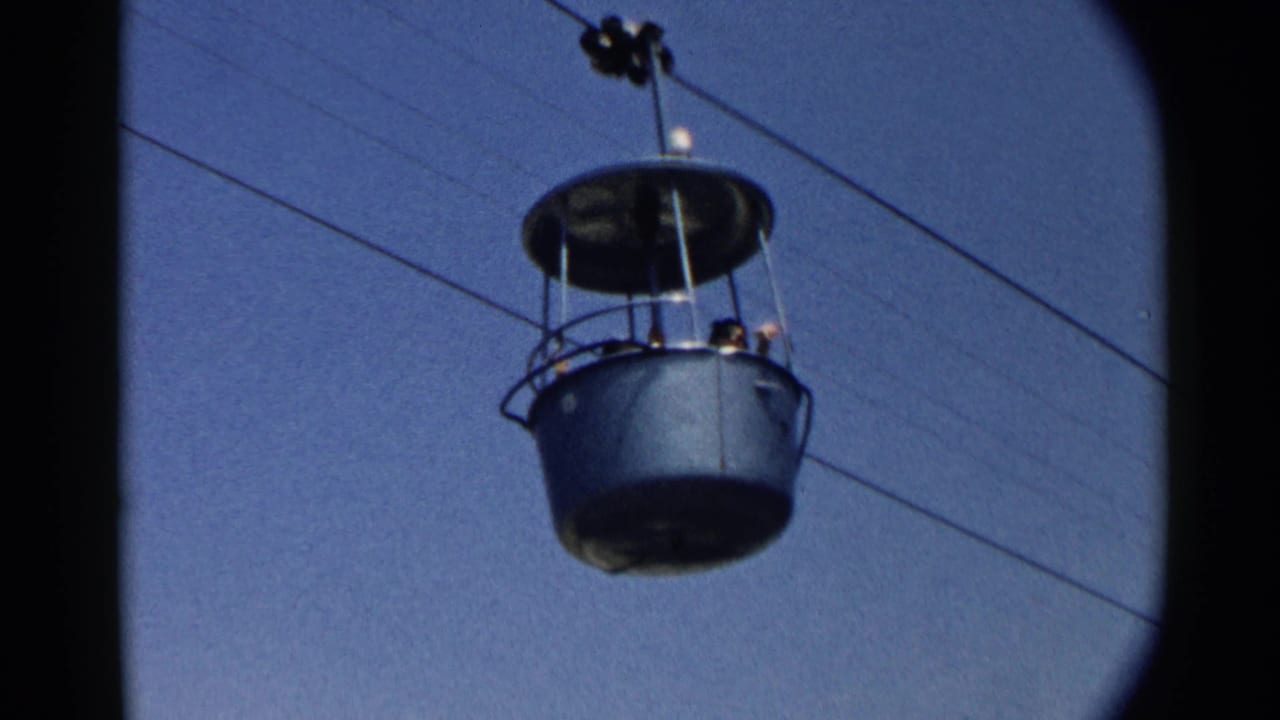 A green overhead cable car cabin with passengers visible, filmed from below against a blue sky in archival 8mm footage.