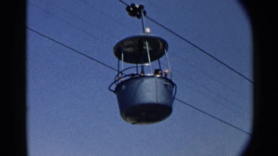 A green overhead cable car cabin with passengers visible, filmed from below against a blue sky in archival 8mm footage.