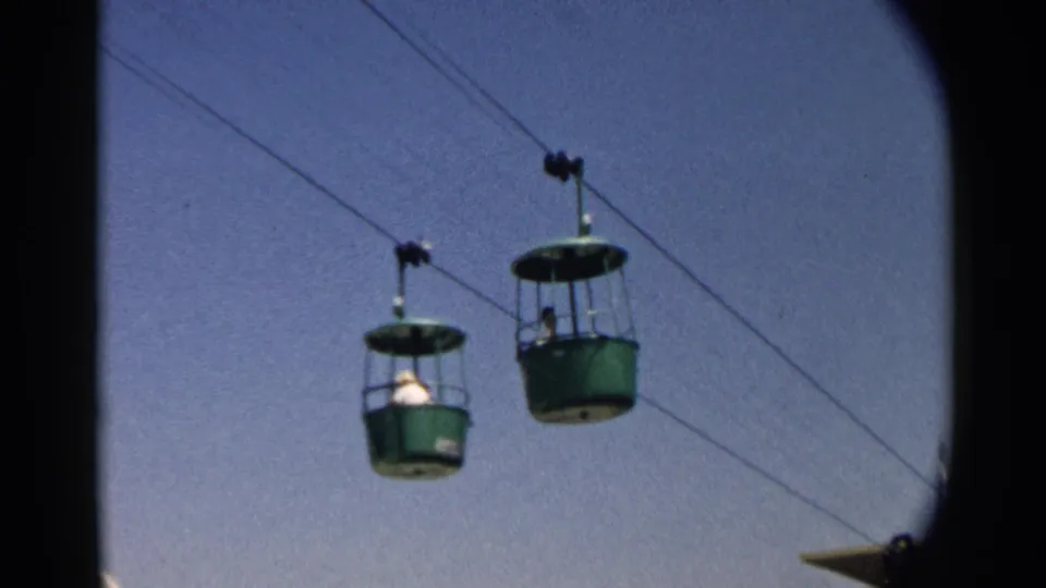 Two green gondola cabins move along suspended wires, with distant passengers visible in the open-air ride.