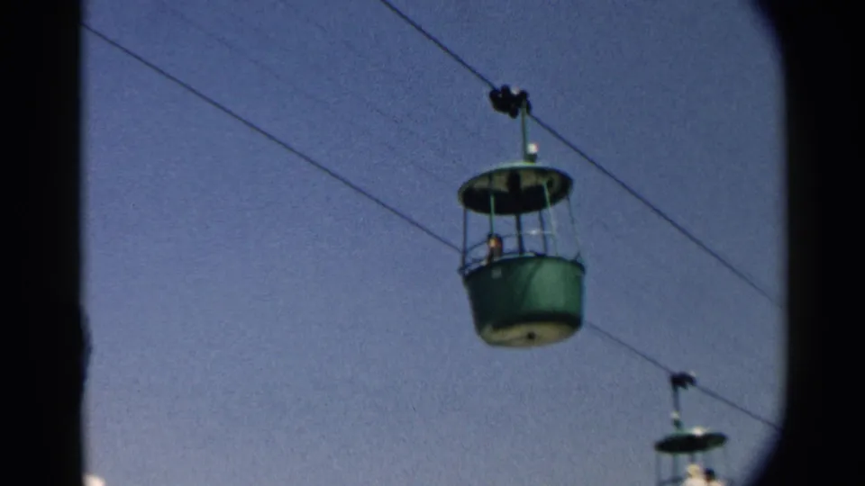 A green cable car cabin passes along diagonal wires, with another smaller cabin lower in the frame and blue sky behind.