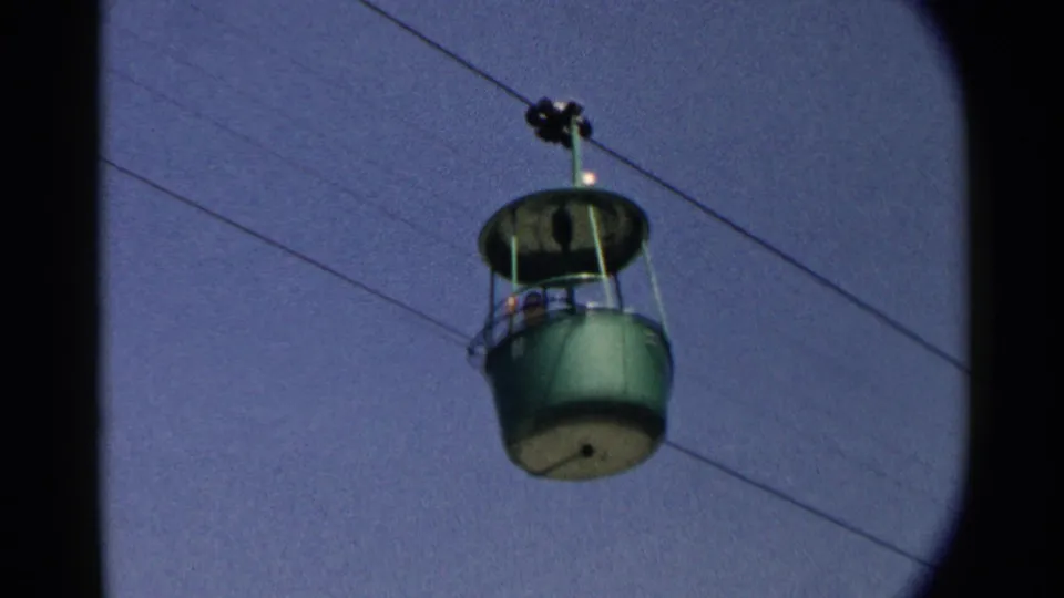 A green overhead gondola cabin appears closer to the camera, carrying visible passengers beneath suspended cables.