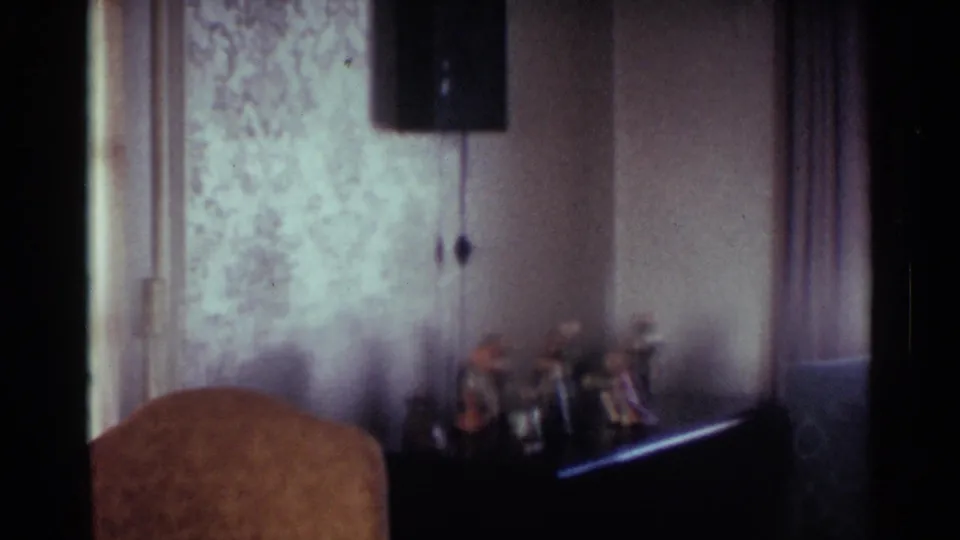 1959 Ohio living room frame showing patterned wallpaper, a wall fixture, drapes, a chair back, and small figurines on a shelf.