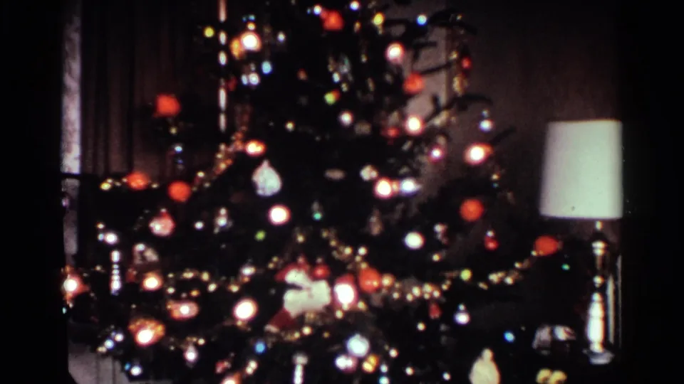 1959 Ohio home-movie frame showing a lit Christmas tree with ornaments and tinsel near a table lamp in a private living room.