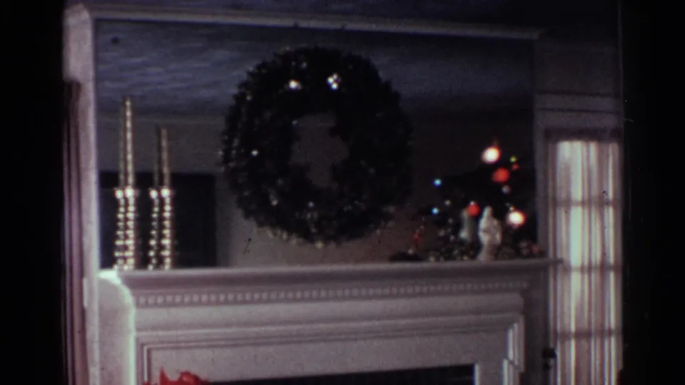 1959 Ohio Christmas interior with a wreath centered above the fireplace, candlesticks on the mantel, and a lit tree near a side window.