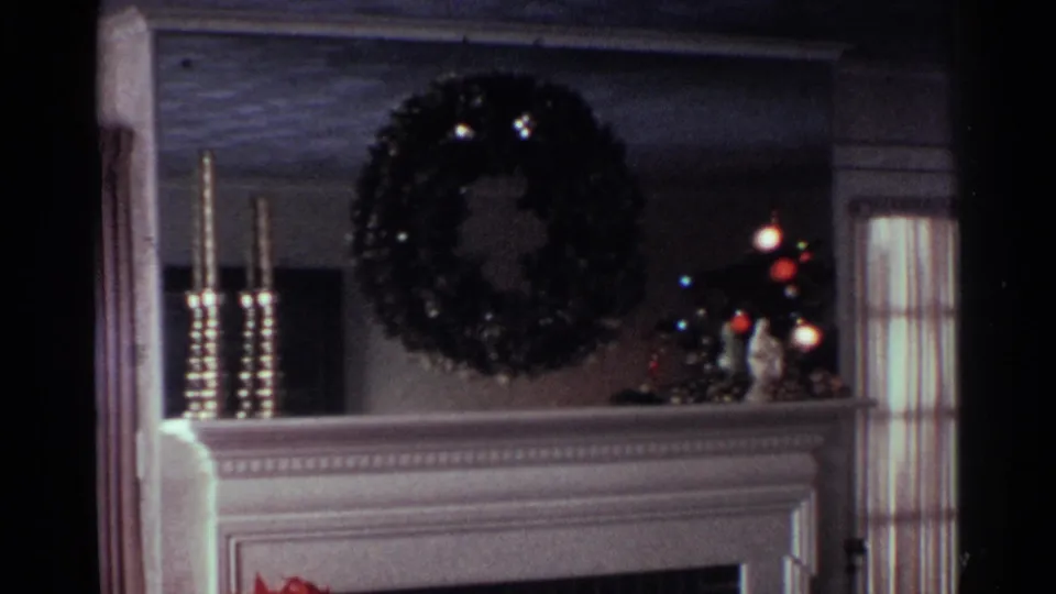 1959 Ohio home-movie frame of a decorated mantel with a dark wreath, tall candlesticks, a small figurine, and a lit Christmas tree.