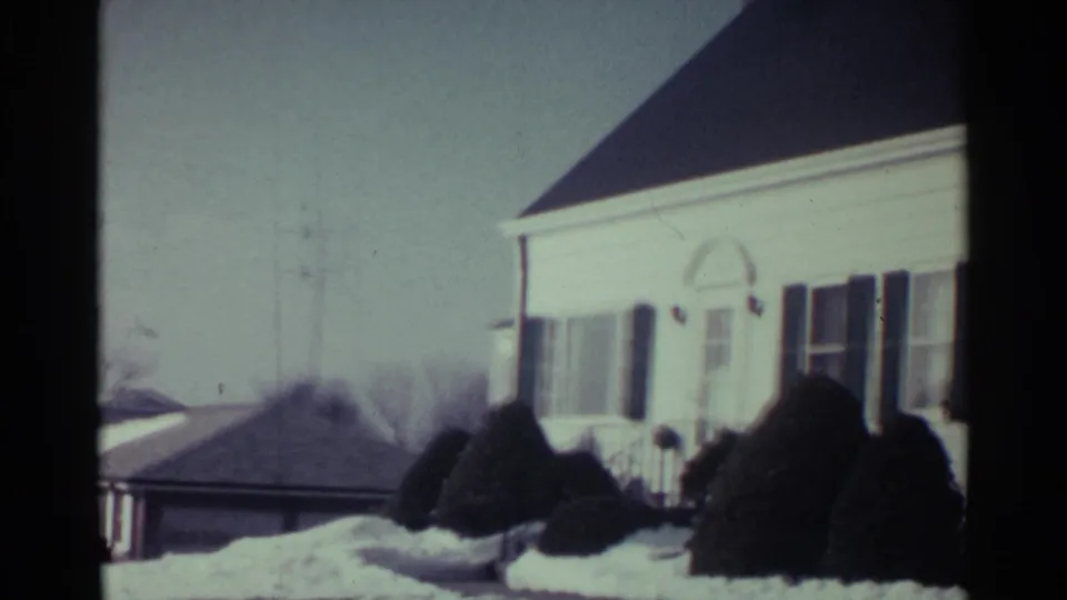 A snow-covered suburban house exterior in Ohio appears with trimmed shrubs, rooflines, and pale winter light in an 8mm frame.