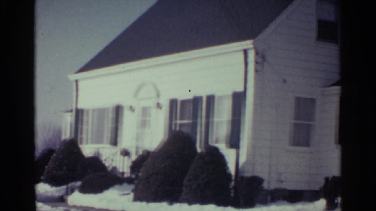 1959 Ohio home-movie frame of a white house exterior with dark shutters, front windows, shrubs, and winter snow.