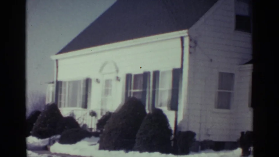 1959 Ohio home-movie frame of a white house exterior with dark shutters, front windows, shrubs, and winter snow.