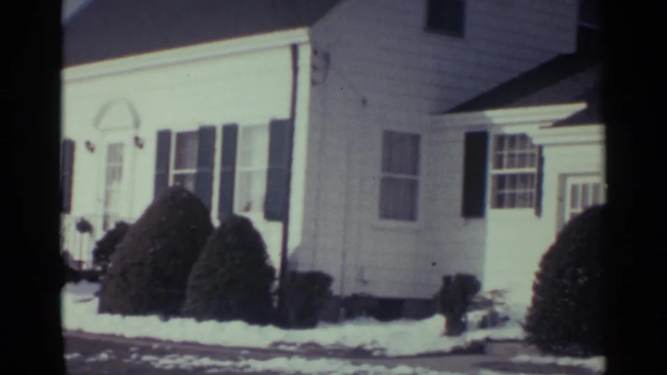 1959 Ohio 8mm view panning across the white house, showing side windows, a door area, shrubs, and snow cover.