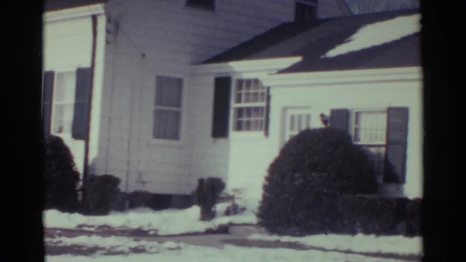 1959 Ohio frame showing the side of a white suburban house, dark shutters, a large rounded shrub, and patches of snow.