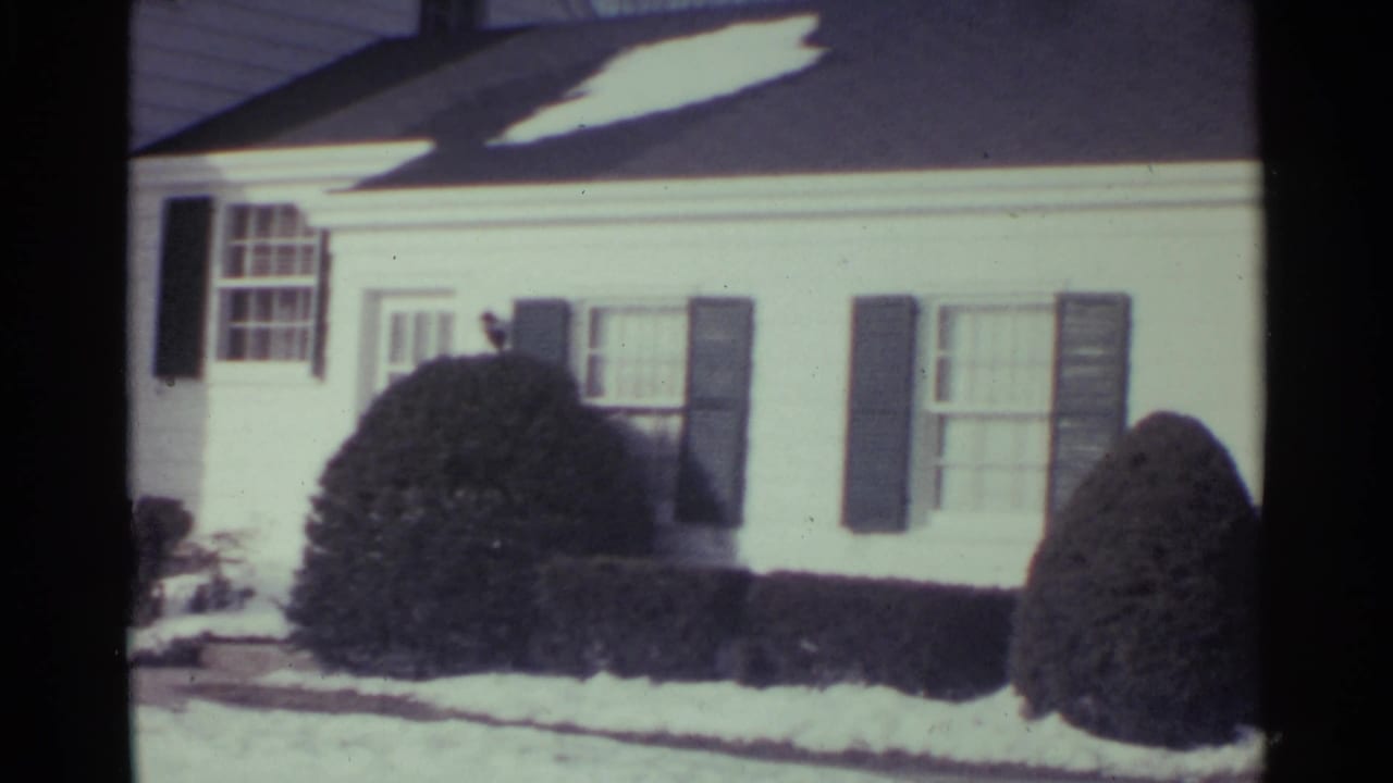 White house facade with shutters, front window, snow, and rounded shrubs in bright sun.