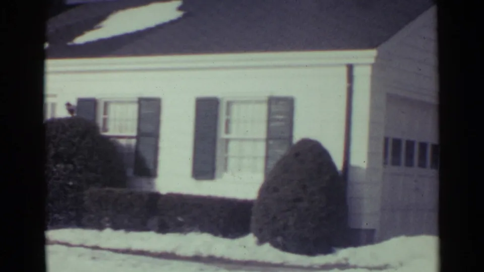 1959 Ohio 8mm frame showing the pan across a white suburban house, dark shutters, shrubs, snow, and the start of an attached garage.