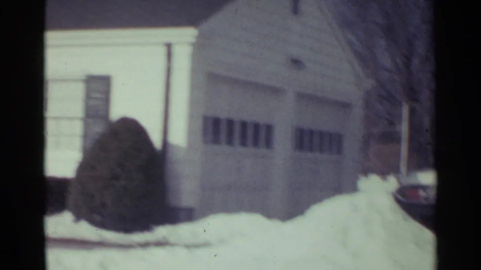 1959 Ohio home-movie frame showing an attached garage, white siding, snow piles, bare branches, and shrubs beside the house.