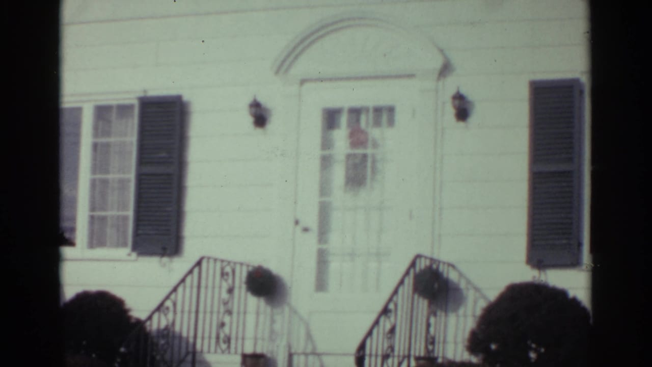White clapboard house front with dark shutters, arched doorway, steps, black railings, and shrubs.