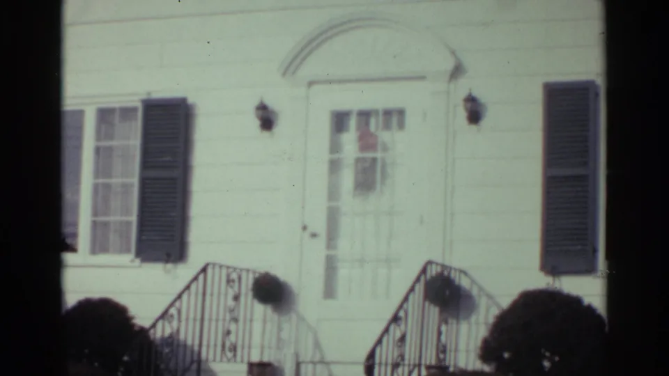 1959 Ohio 8mm frame showing a white residential facade with dark shutters, arched entry trim, metal stair railings, and trimmed shrubs.