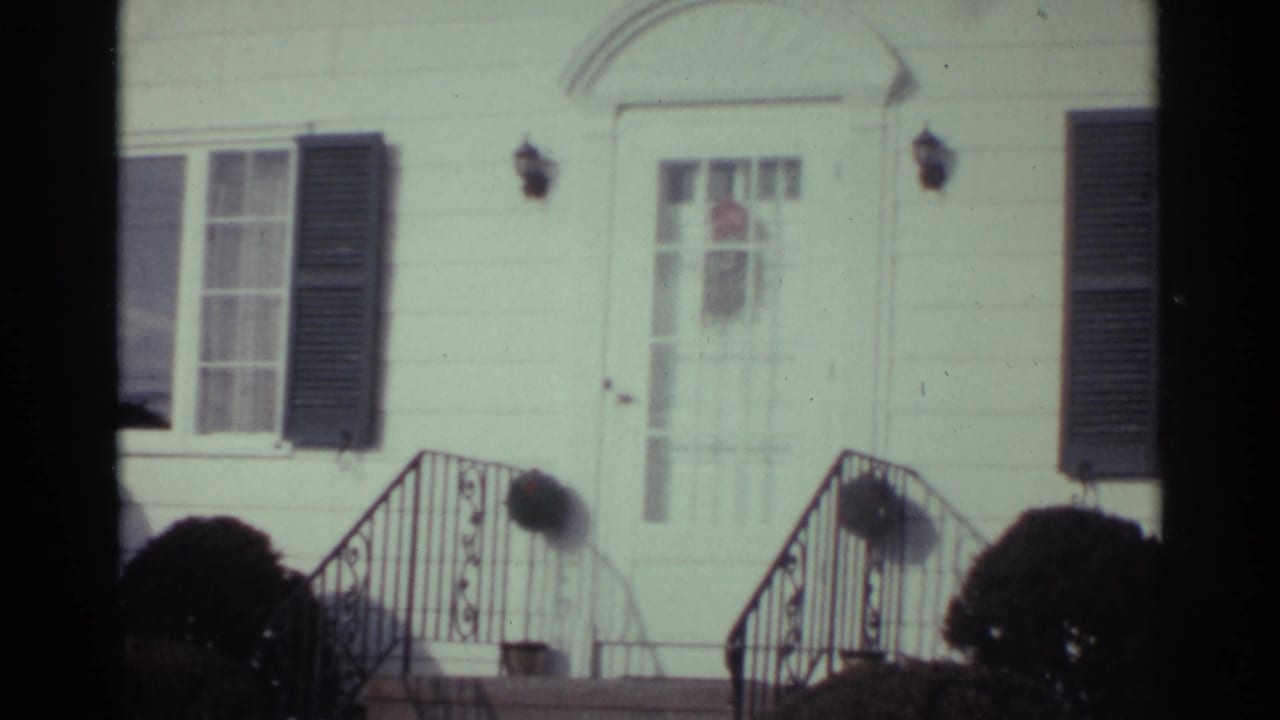 1959 Ohio home-movie frame centered on a white house entrance with dark shutters, porch lamps, black railings, and bushes near the steps.