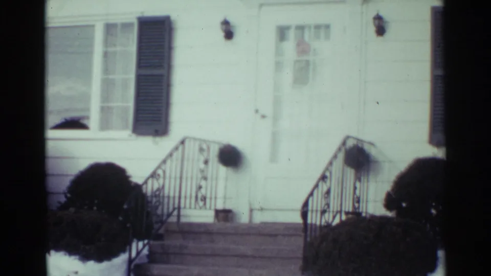 1959 Ohio 8mm frame with a slightly shifted view of the white house front, showing the entry, windows, shutter, stair railings, and clipped shrubs.