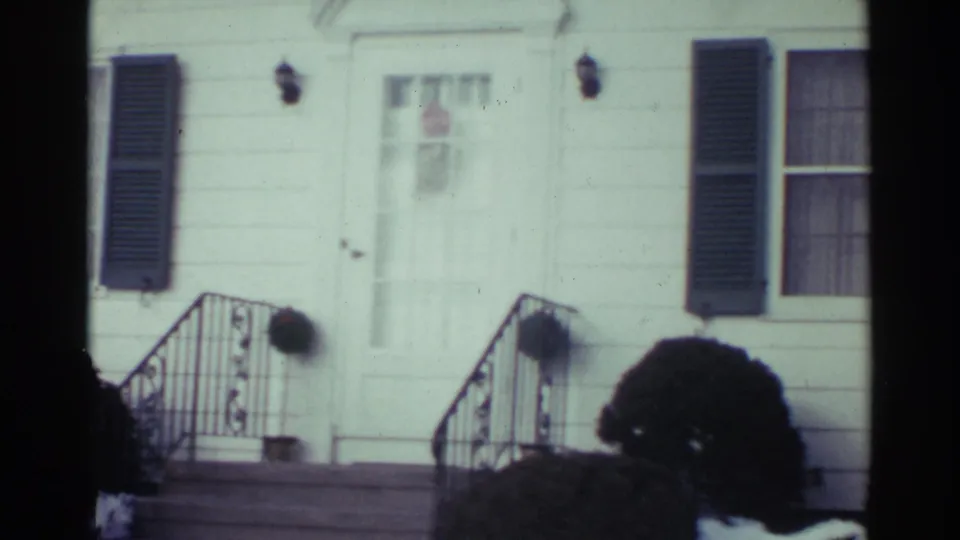 1959 Ohio frame of a white clapboard house front with arched doorway trim, dark shutters, black railings, porch lamps, and bushes by the steps.