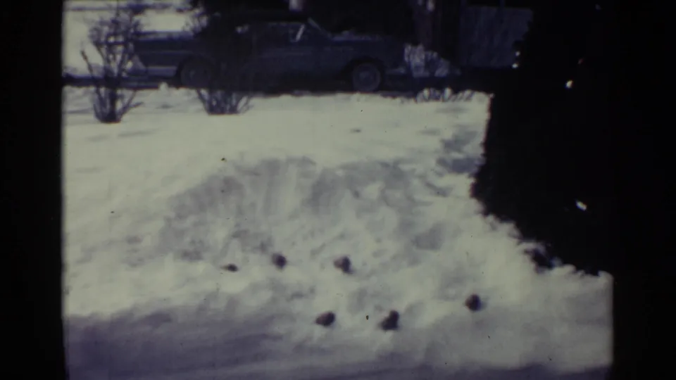 1959 Ohio home-movie frame of a snowy residential exterior with a parked car beyond the snow.
