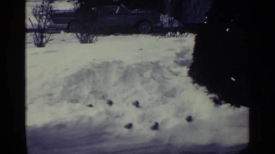 1959 Ohio home-movie frame showing piled snow, bare shrubs, and a parked car beyond the yard.