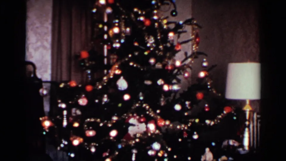 1959 Ohio home-movie frame showing a tall Christmas tree with ornaments, lights, garlands, wallpaper, curtains, and a nearby lamp.