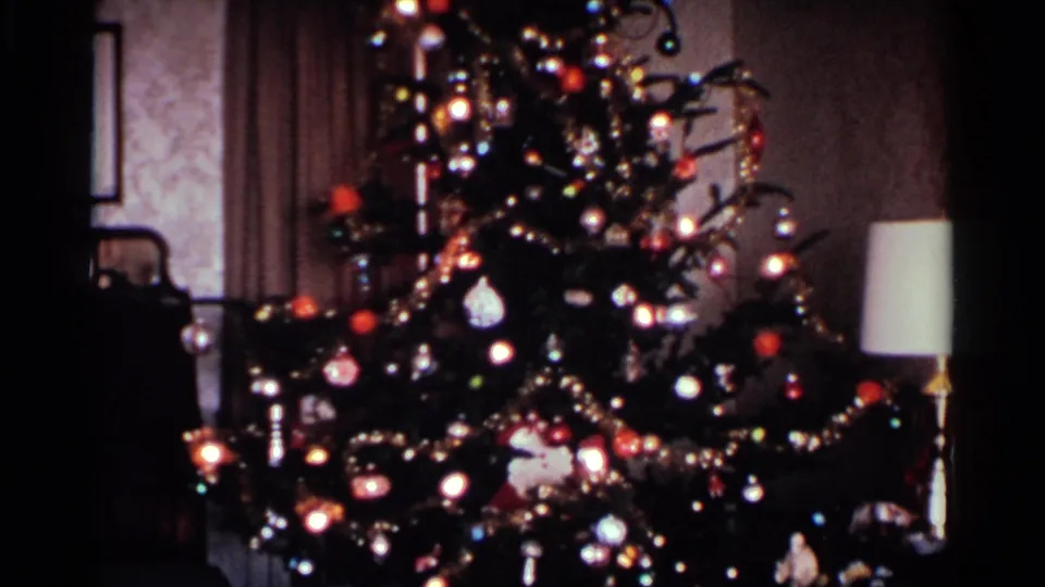 1959 Ohio home interior frame showing the decorated tree closer in view, with bright bulbs, ornaments, garlands, wallpaper, and a lamp at right.