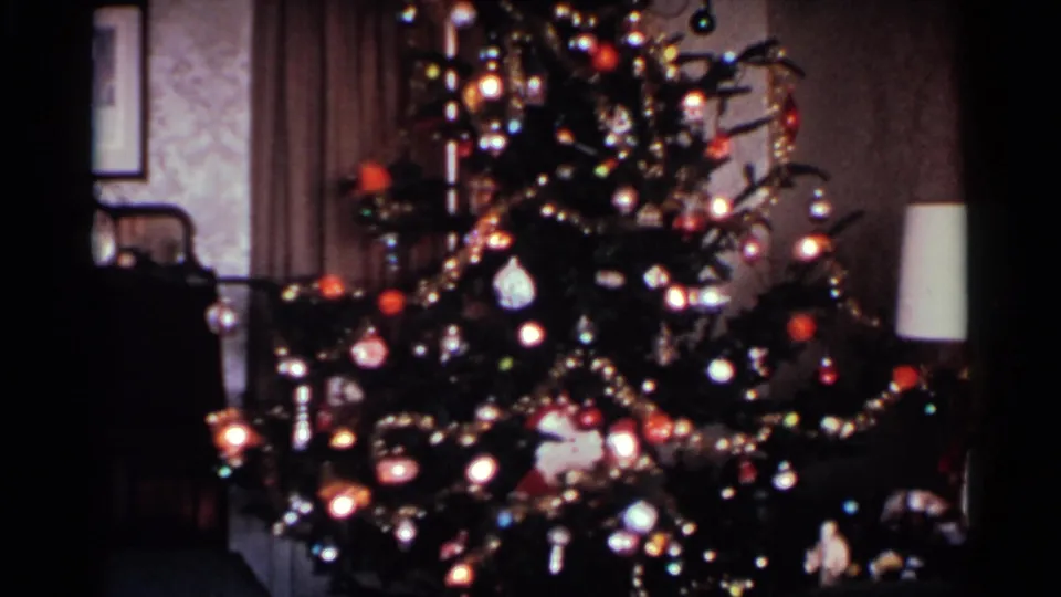 1959 Ohio frame of a tall decorated Christmas tree in a private living room, with lights, ornaments, garlands, a lamp, wallpaper, and curtains.