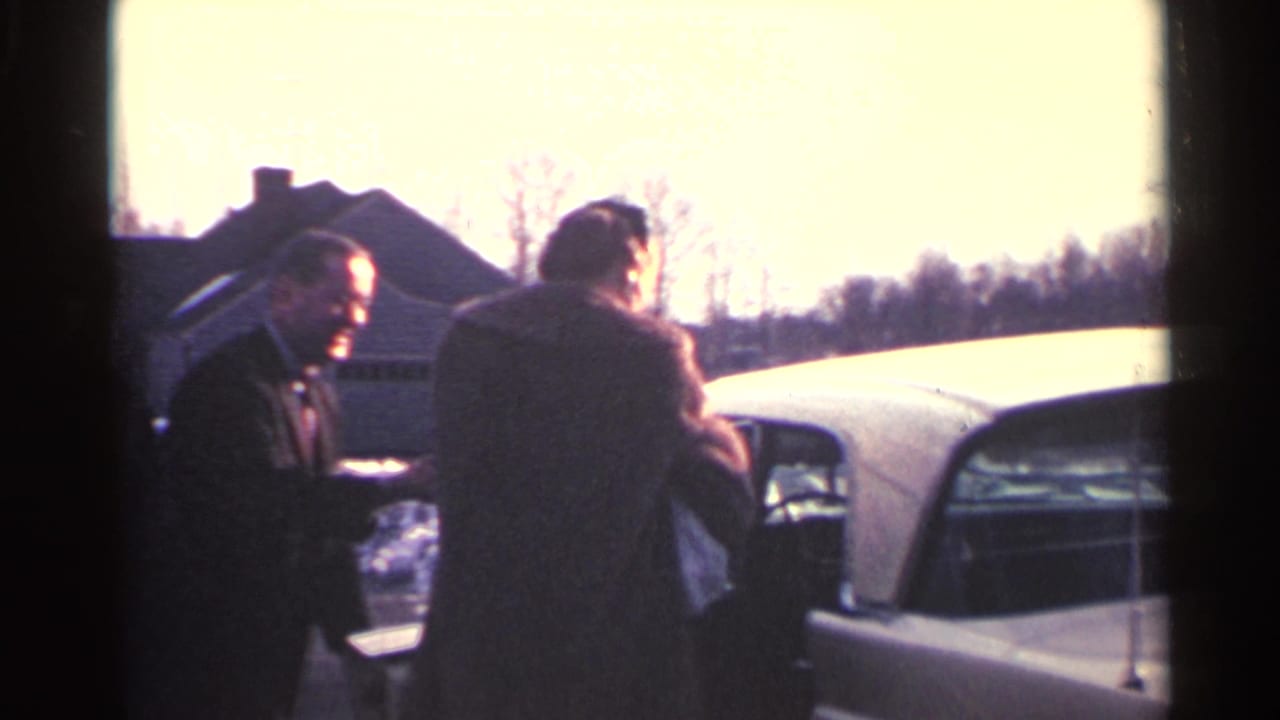 In Ohio home-movie footage from 1959, two adults in coats stand by a white sedan with houses and bare trees behind them.