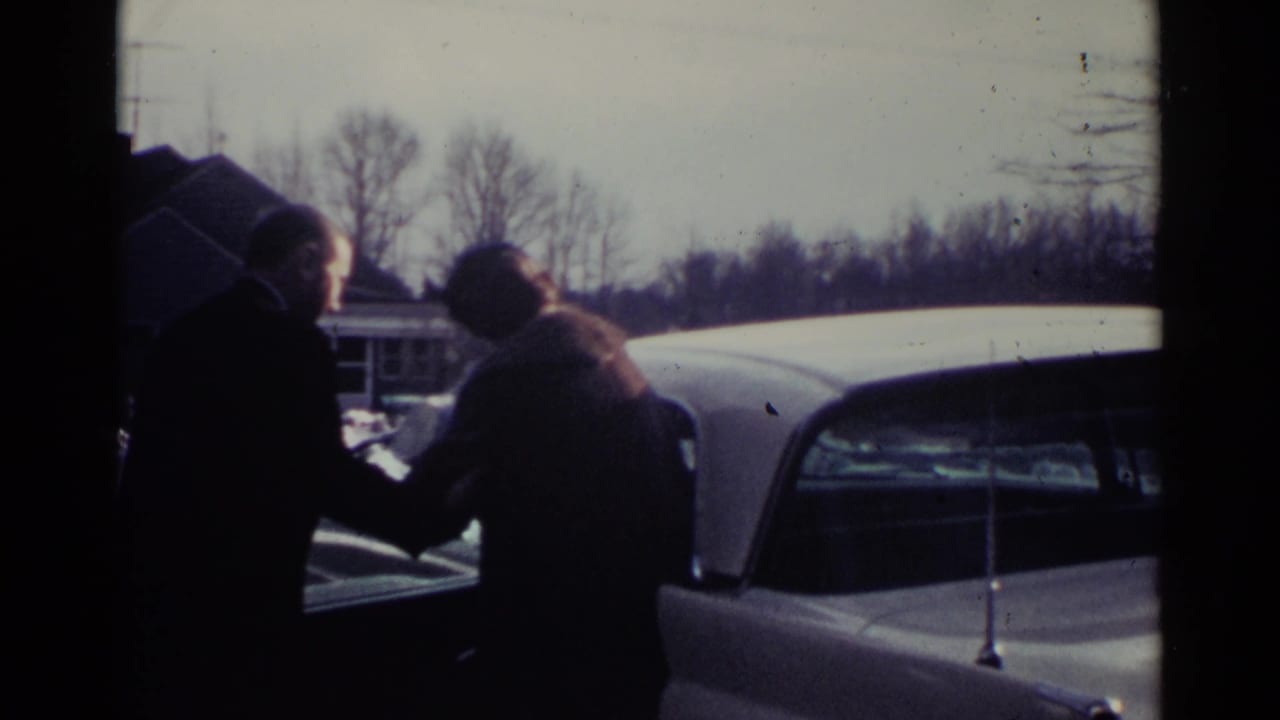 A man in a dark coat appears to assist a woman beside a white vintage car in the 1959 Ohio suburban scene.