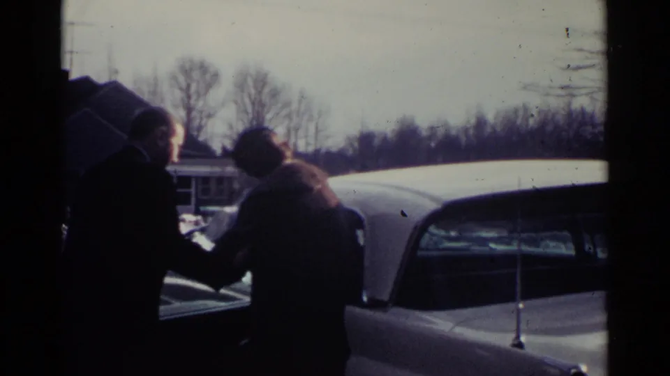 A man in a dark coat appears to assist a woman beside a white vintage car in the 1959 Ohio suburban scene.