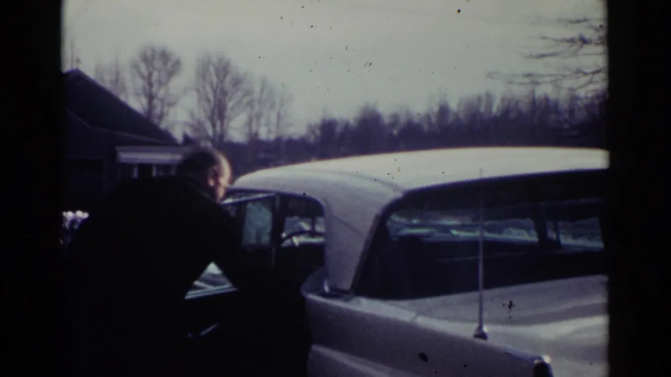 The man bends near the open door of the white car, with winter trees and a house visible in the Ohio home-movie frame.
