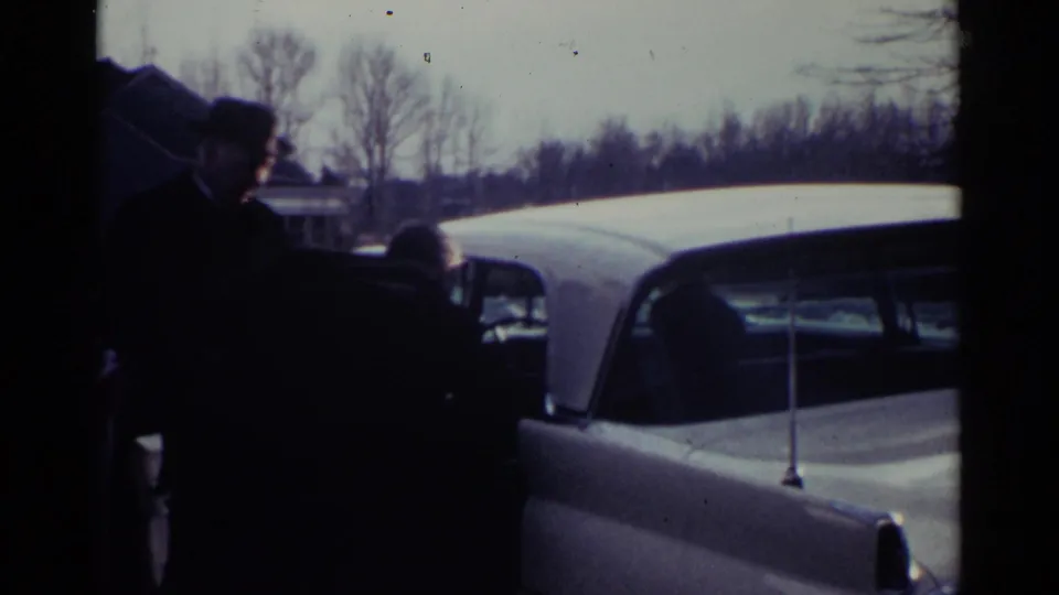 Two adults cluster at the open door of a white sedan, completing the brief assisted-entry moment in 1959 Ohio footage.