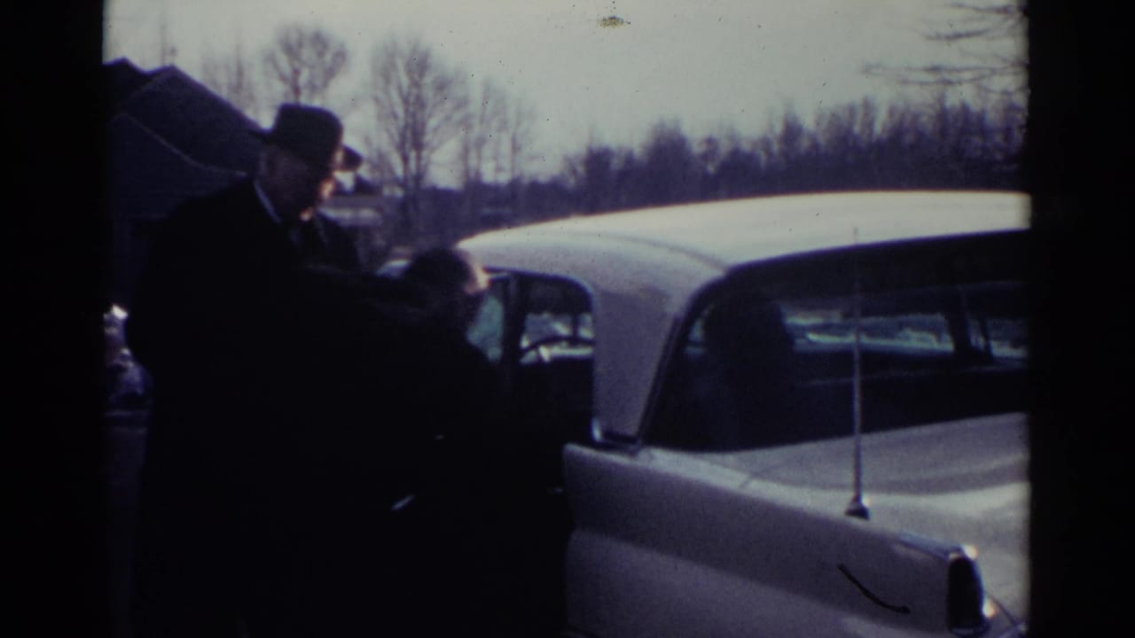 Man in a dark suit and fedora standing beside a white vintage car outdoors.
