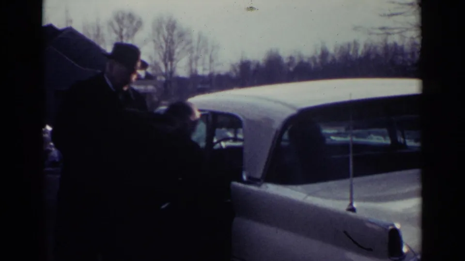 In Ohio 1959 home-movie footage, a suited man in a fedora stands near a white vintage car in a suburban driveway or roadside setting.
