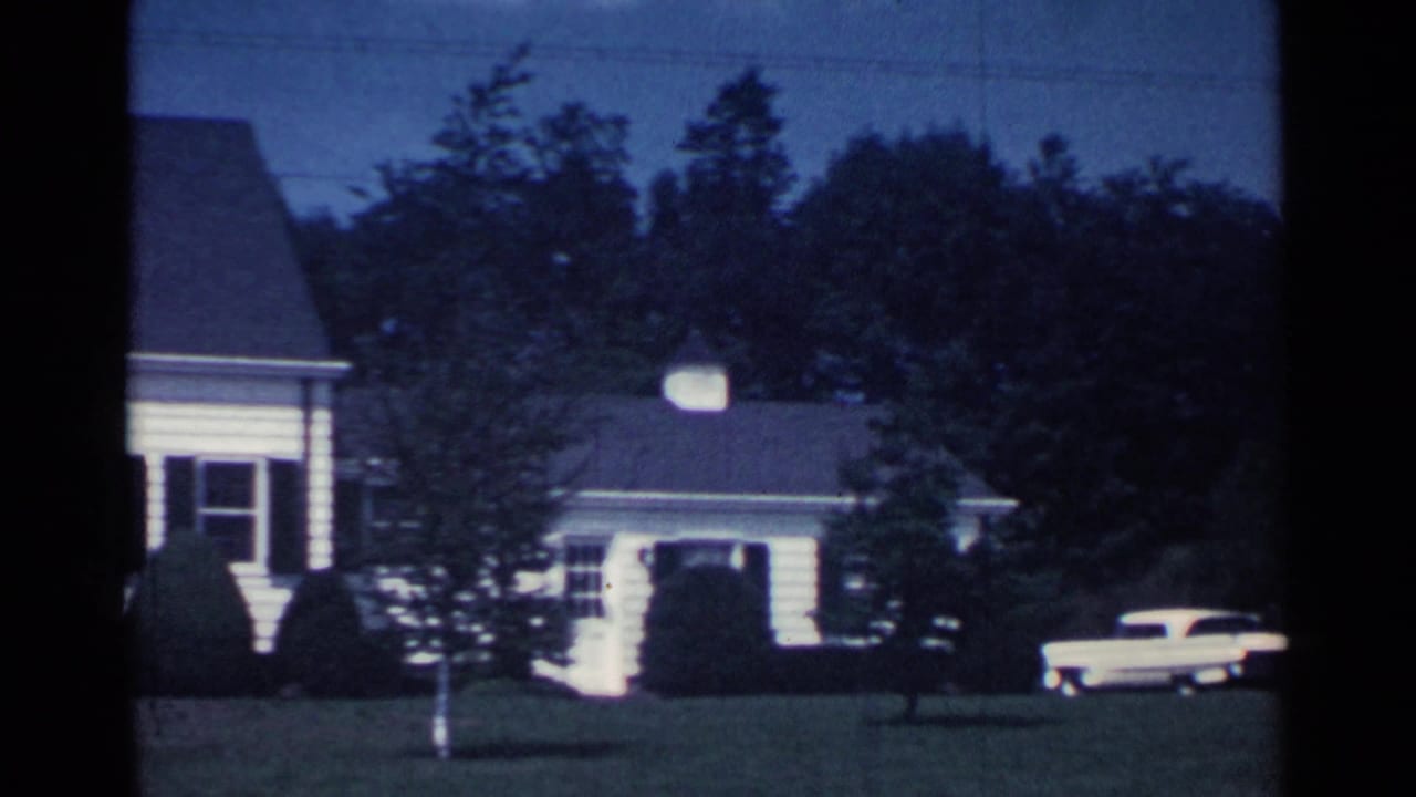 White suburban house, lawn, trees, and a pale vehicle at dusk.