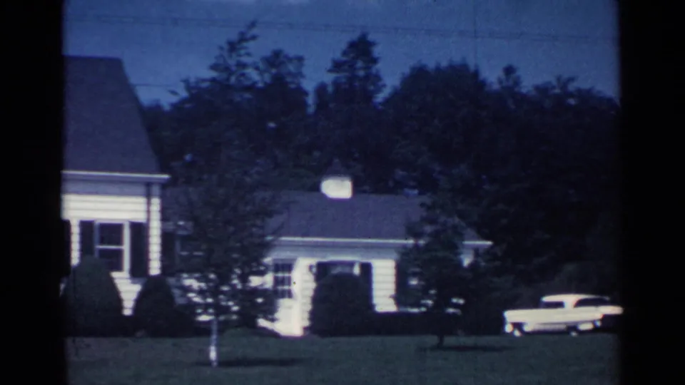 1959 Ohio 8mm frame showing a white suburban house, mown lawn, trees, and a pale period vehicle in blue dusk light.