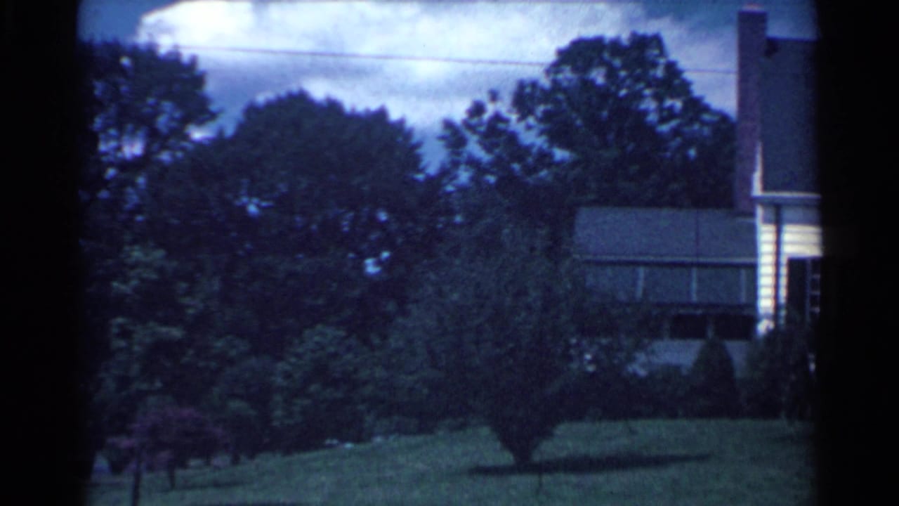 1959 Ohio 8mm frame showing a white suburban house partly obscured by trees, with lawn, shrubs, and cloudy sky.