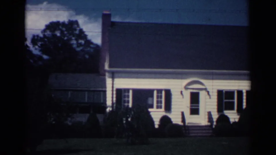 1959 Ohio 8mm frame showing the front of a white suburban home with shutters, entry steps, shrubs, and side porch area.