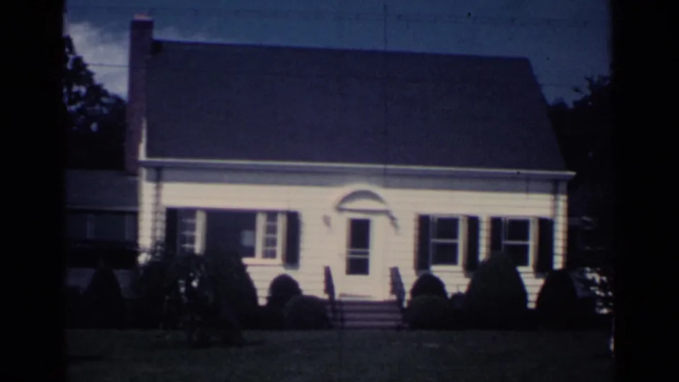 1959 Ohio home-movie frame of a white suburban house front, with dark shutters, chimney, shrubs, lawn, and trees.