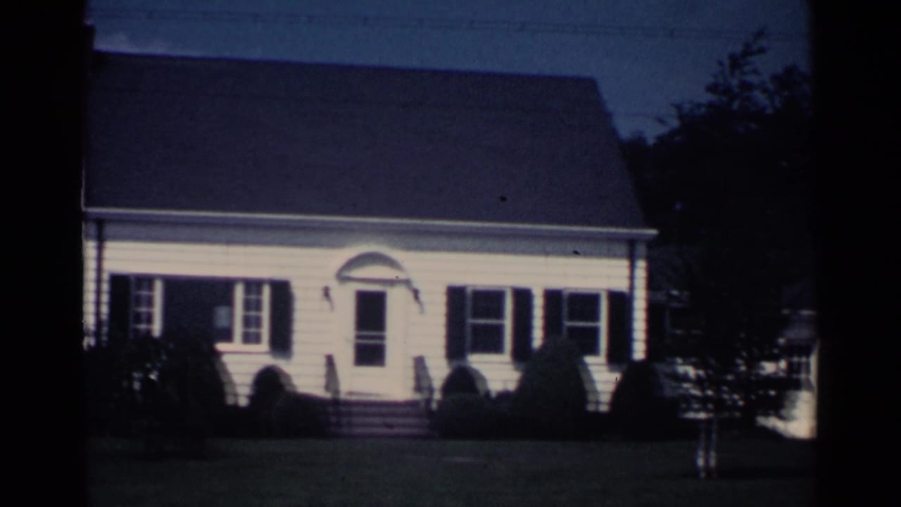 White clapboard house with dark shutters, front steps, lawn, shrubs, and trees.