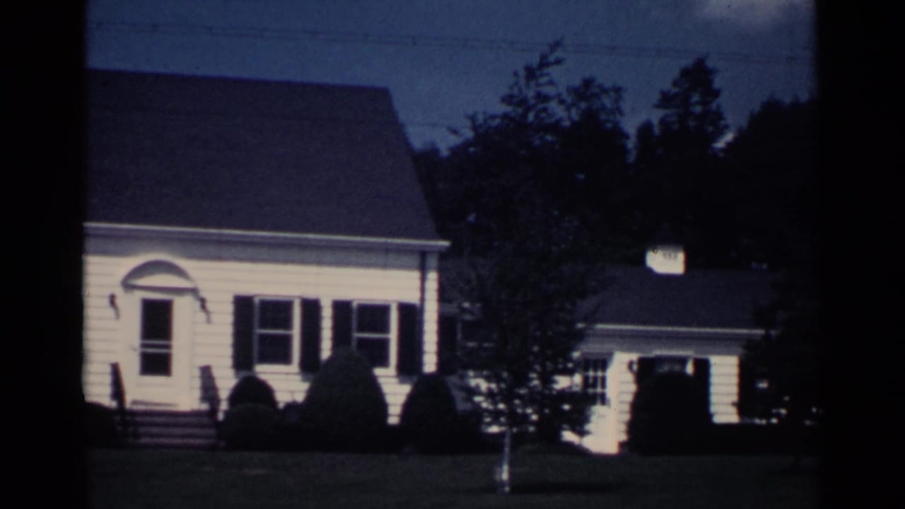 House facade with dark shutters, front doorway, lawn, shrubs, trees, and garage at right.