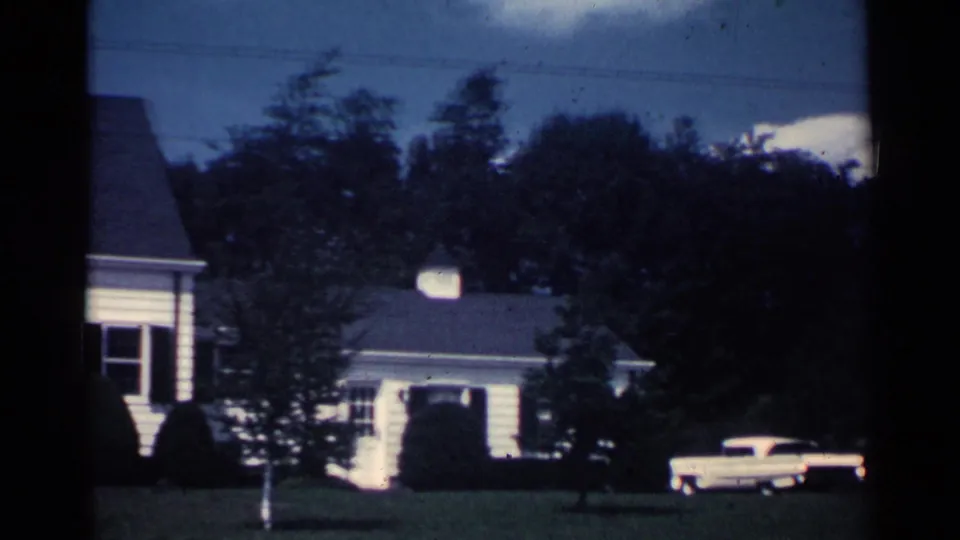 1959 Ohio home-movie frame with part of the house, a detached garage, and a white 1950s sedan visible beside the yard.