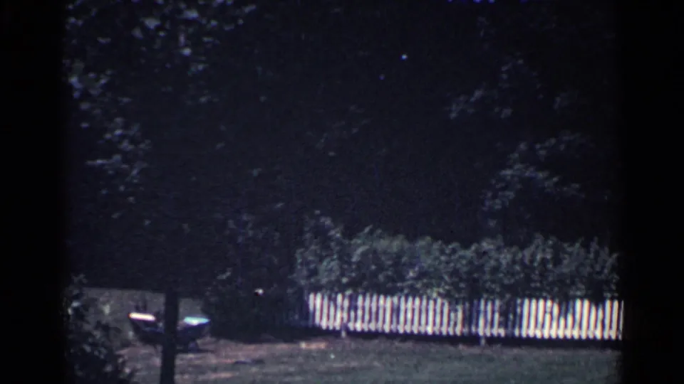 Final 1959 Ohio frame showing a darker section of the yard with trees, lawn, a white picket fence, and shadowed structures.
