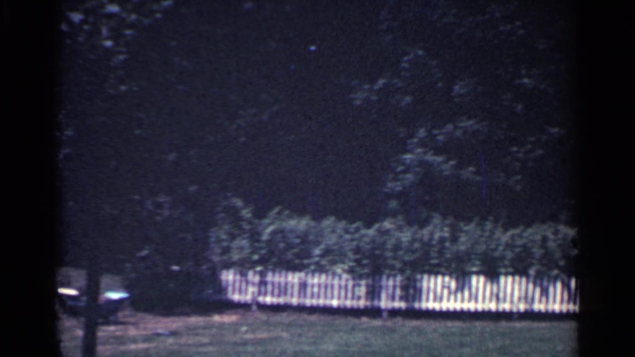 1959 Ohio 8mm frame showing a white picket fence along a green yard with shrubs and a dark wooded backdrop.