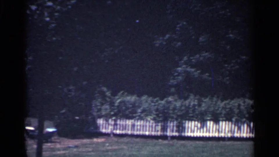 1959 Ohio 8mm frame showing a white picket fence along a green yard with shrubs and a dark wooded backdrop.